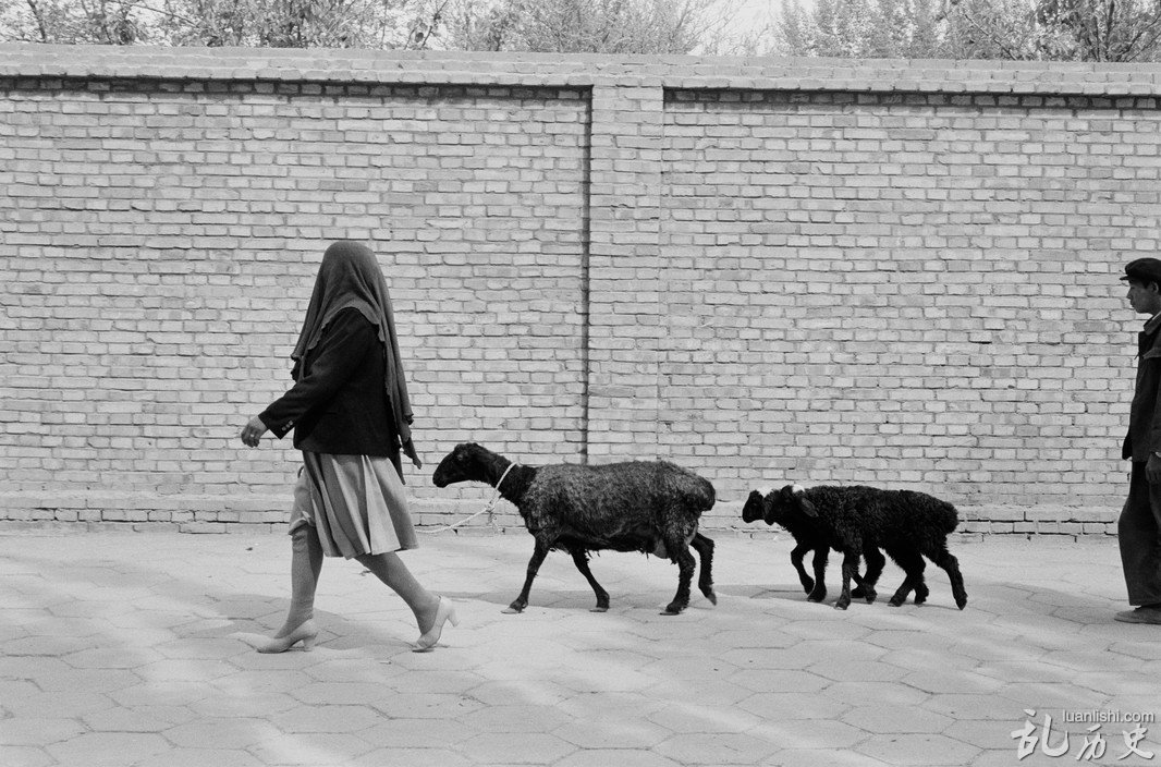 1989年，中国新疆喀什，戴着头巾的维族妇女牵着羊儿从街上走过。